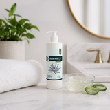 A bottle of Aloe Vera gel treatment stands on a marble countertop in a bathroom setting, with a rolled white towel, a small glass bowl of clear gel, aloe vera slices, a potted plant, and a round mirror in the background.