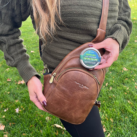 Person holding a brown leather bag and Zatural social gummies outdoors on grass